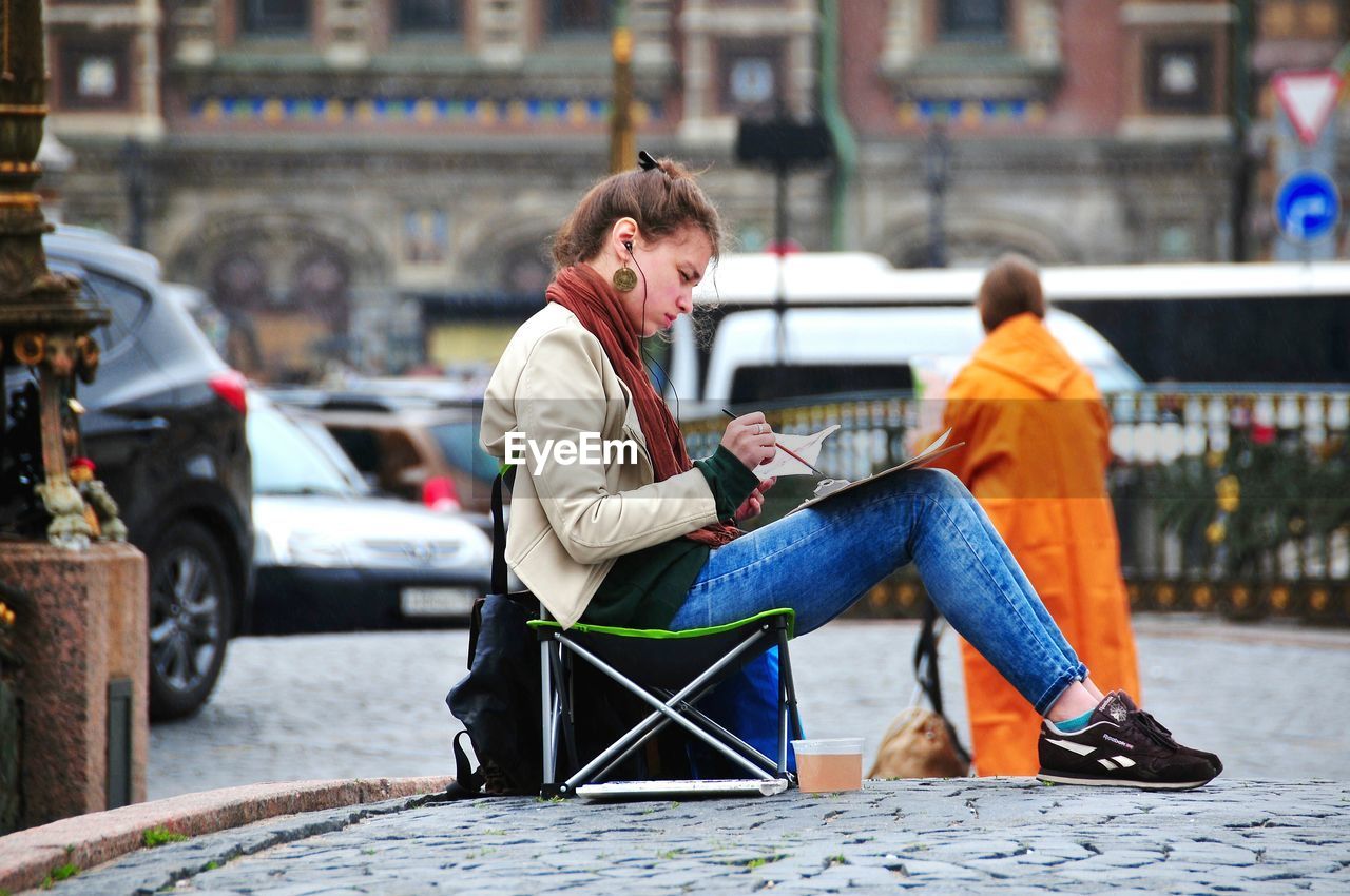 Woman painting while sitting on cobbled street in city