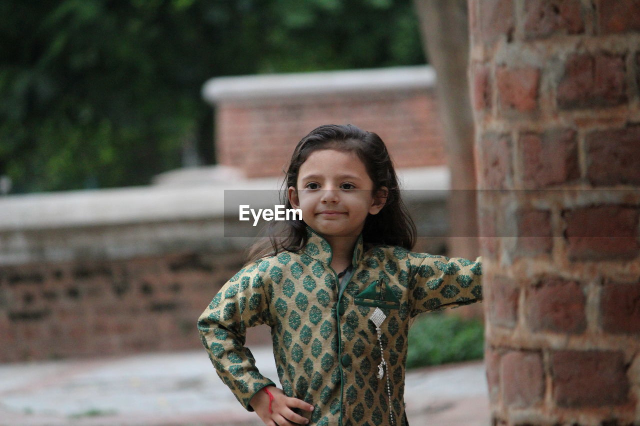 PORTRAIT OF A SMILING GIRL STANDING AGAINST BRICK WALL