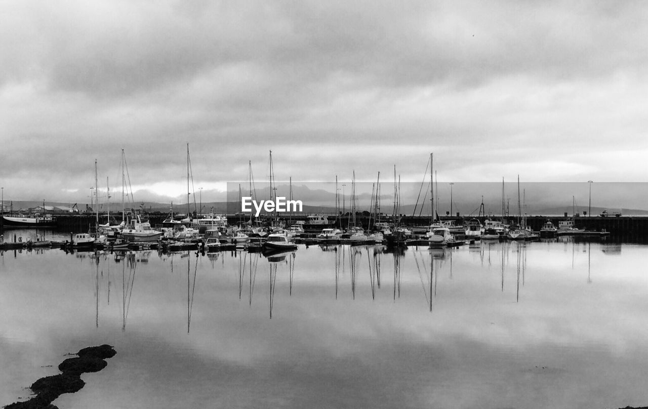 Sailboats moored at harbor against cloudy sky