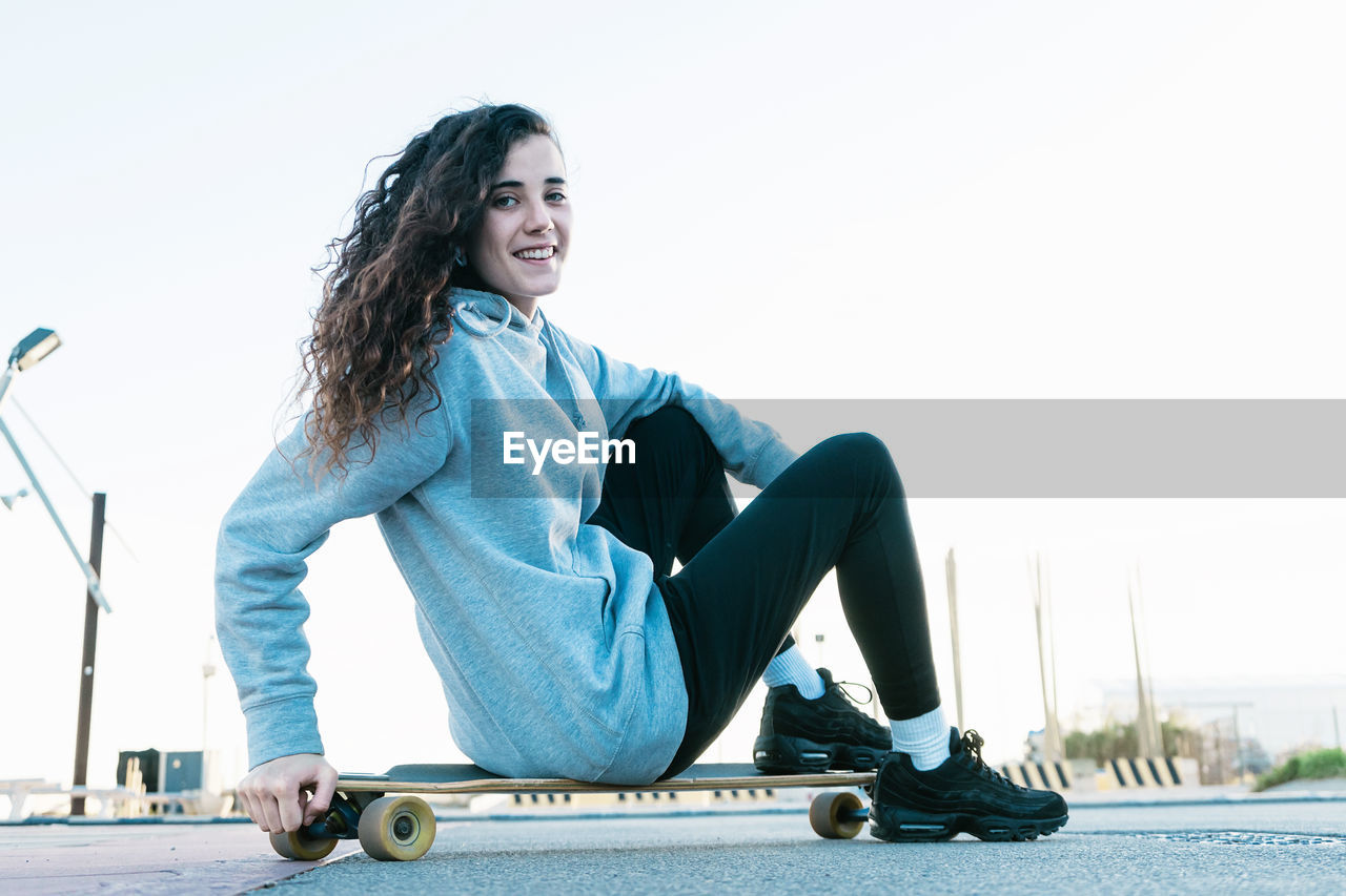 Smiling teenage girl sitting on skateboard against sky