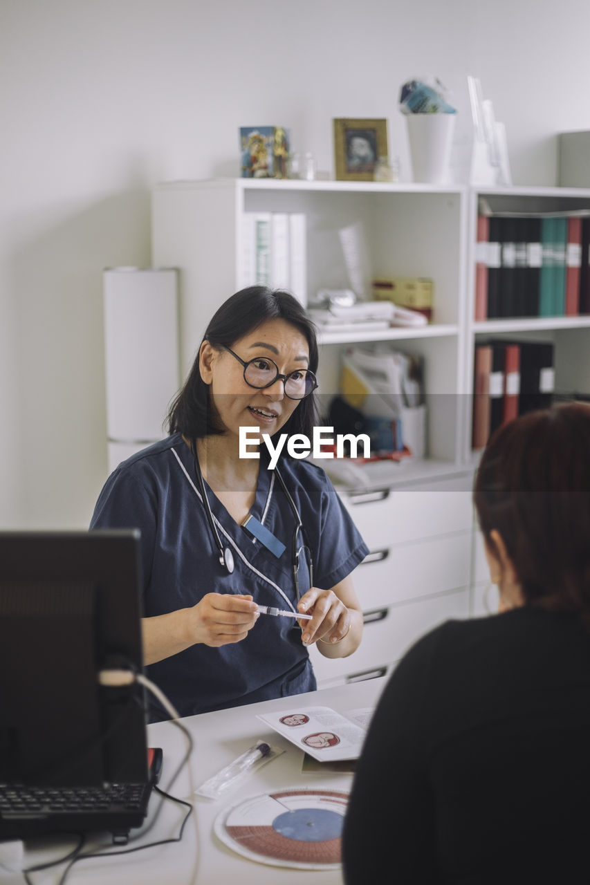 Female doctor explaining ivf syringe to young patient while sitting at desk in medical clinic