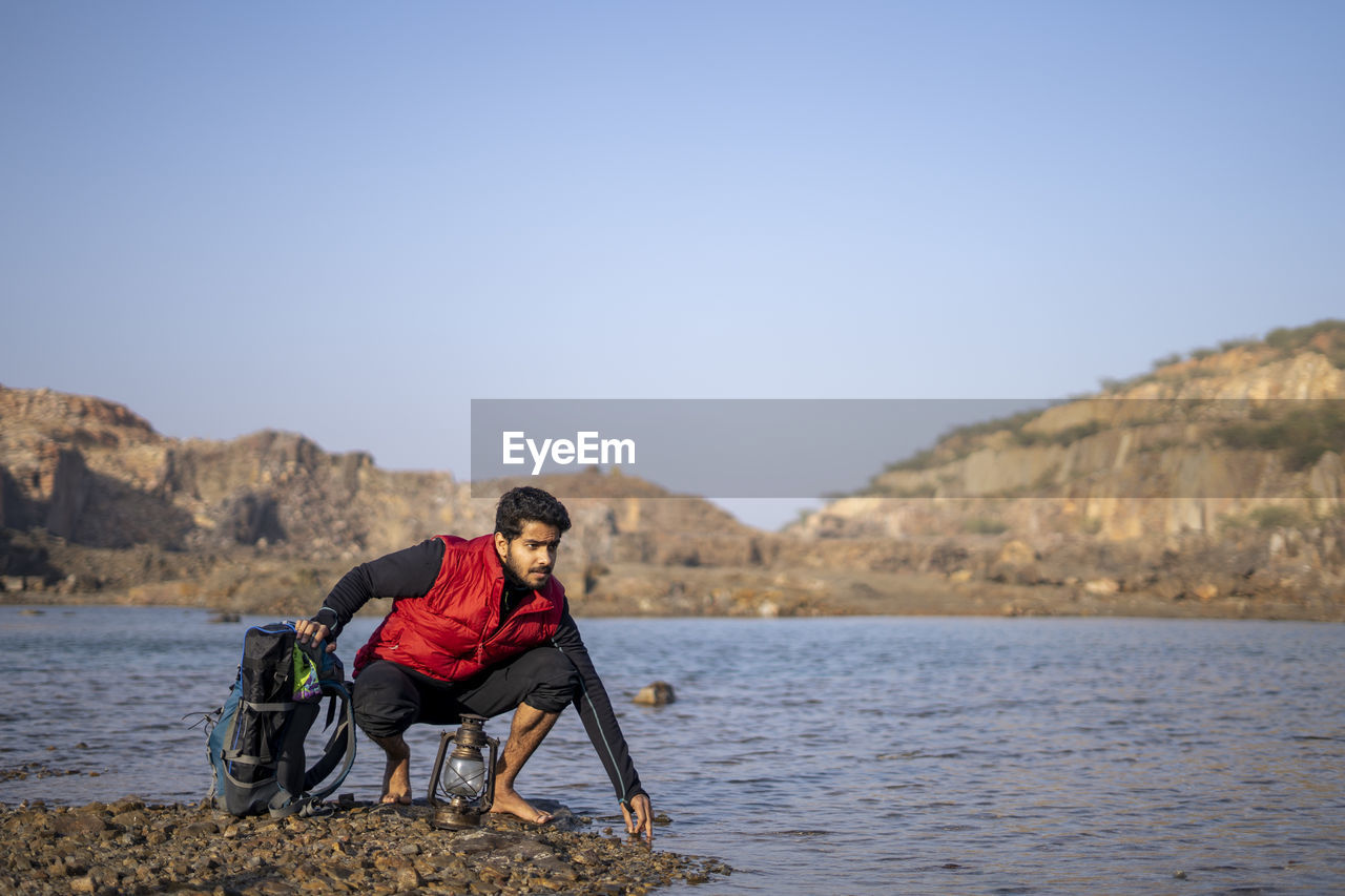 Young indian traveler getting ready for camping in the mountains, sitting near a lake with his pack.