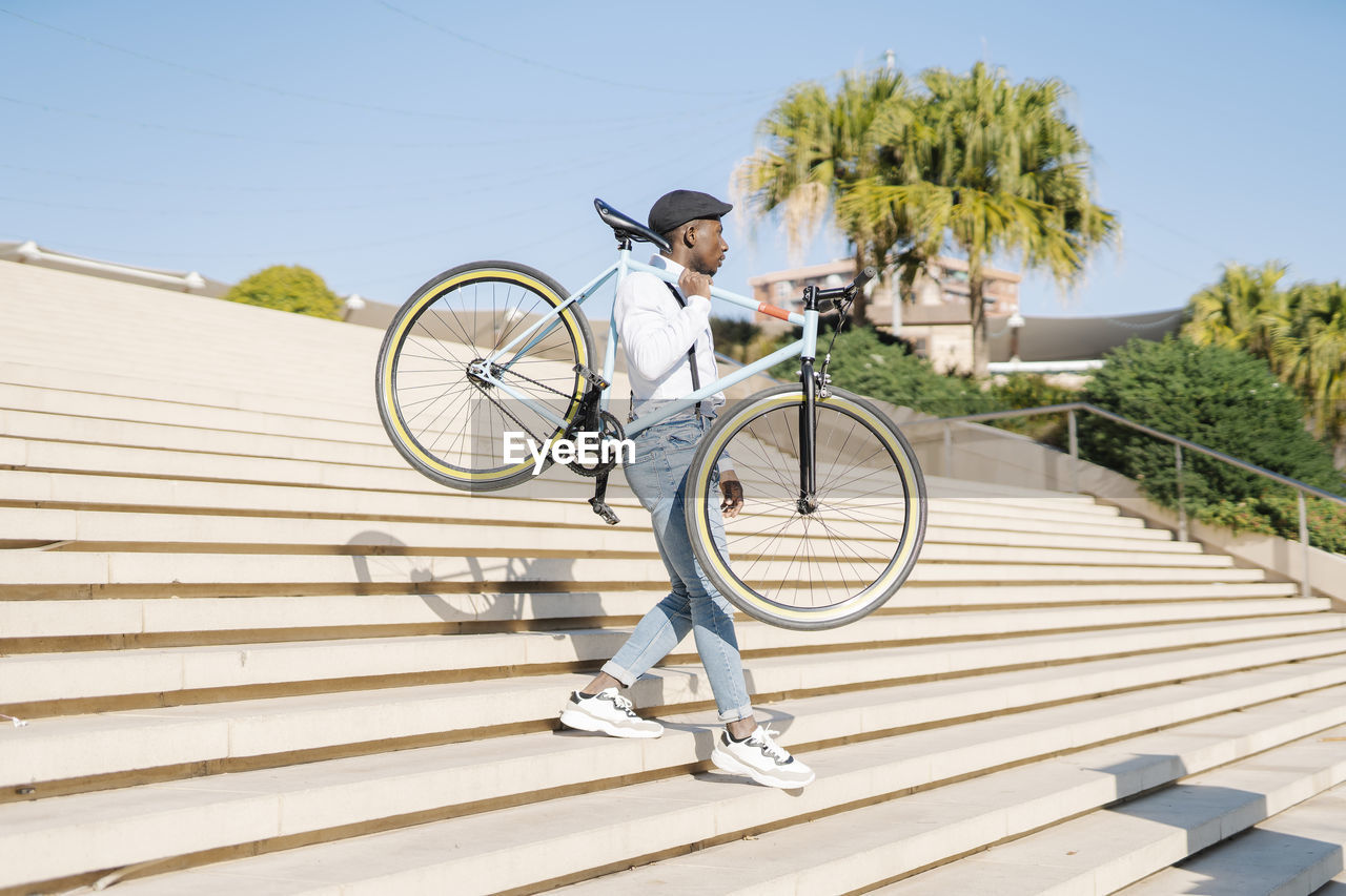 Young man carrying bicycle and moving down staircase on sunny day
