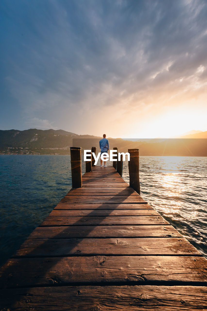 Young man on a wooden jetty on lake orta as he watches the sun set behind the mountains at sunset