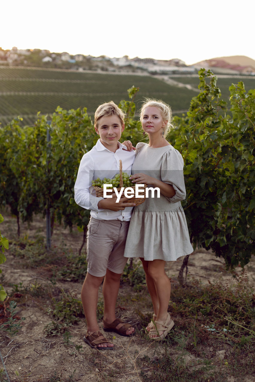 Mother and her teenage son standing in a vineyard with a wicker basket and green grapes in nature