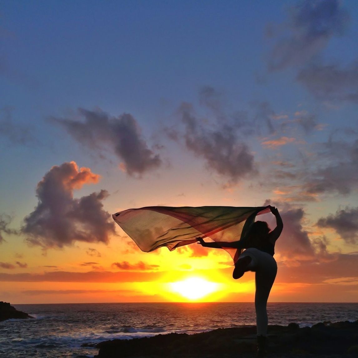 Rear view of woman with scarf standing at beach against sky during sunset