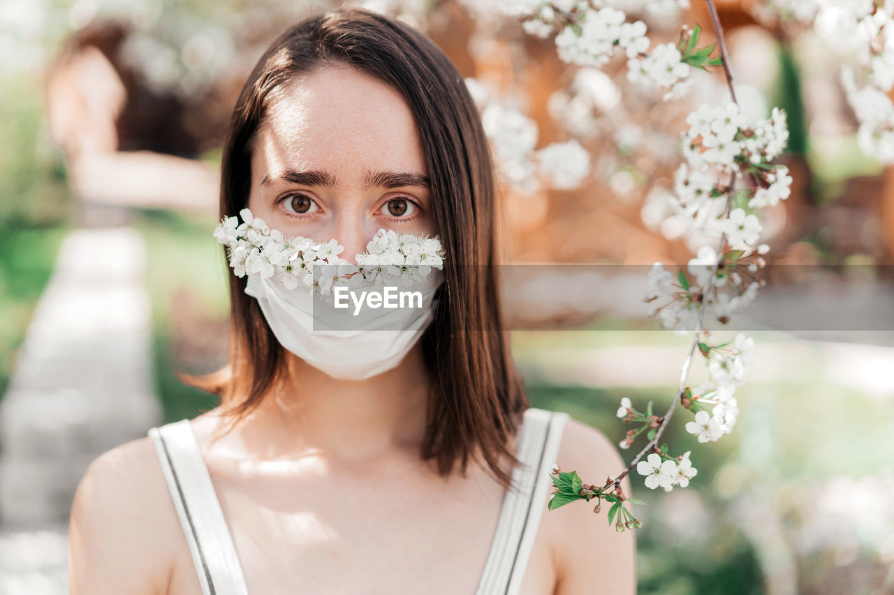 Portrait of sad young woman in protective medical face mask with flowers near blooming tree.