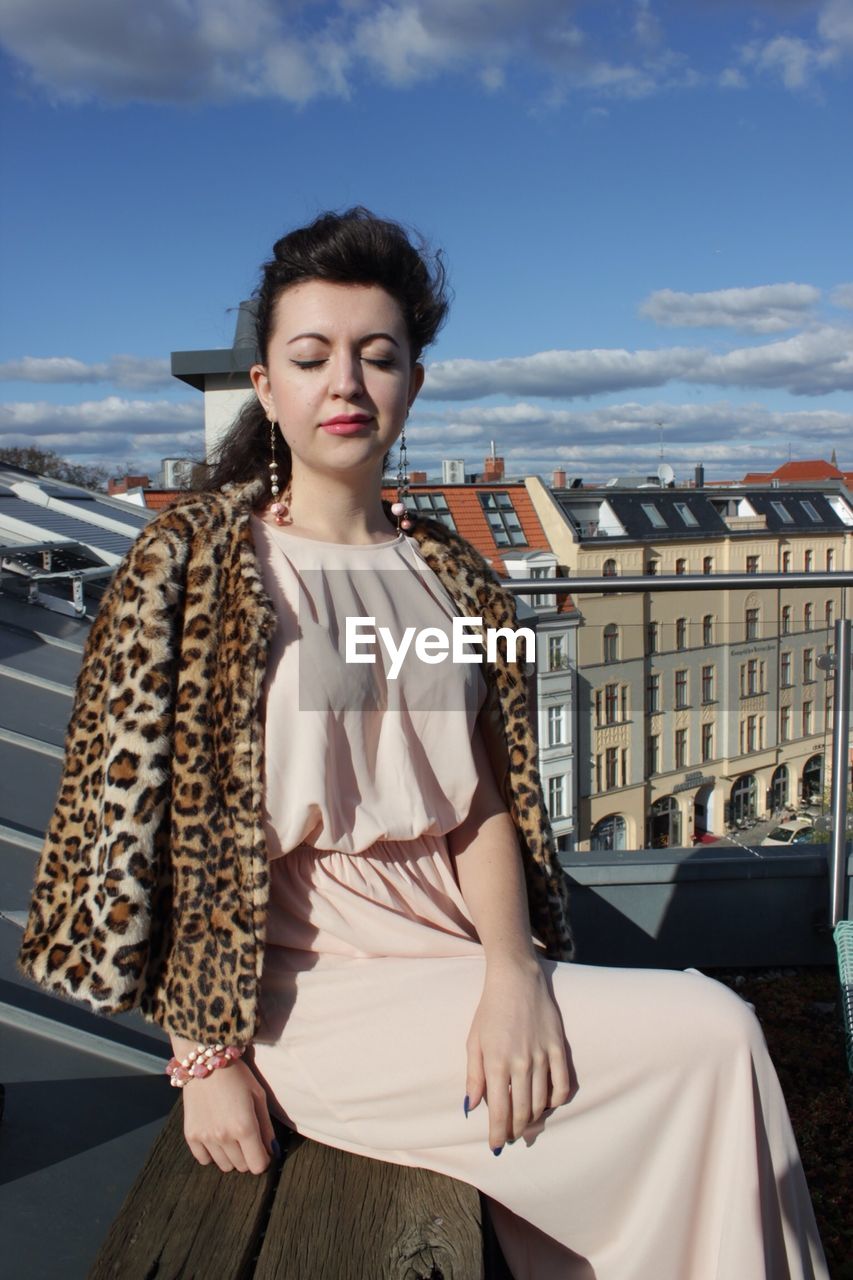 Beautiful woman sitting with eyes closed on building terrace against sky