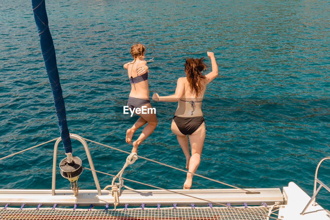 Two women friends jump off catamaran sailing yacht into blue sea in the mediterranean during summer.