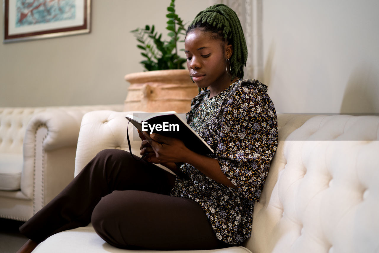 Side view of focused african american female with braids reading interesting book while resting on couch in modern living room at home