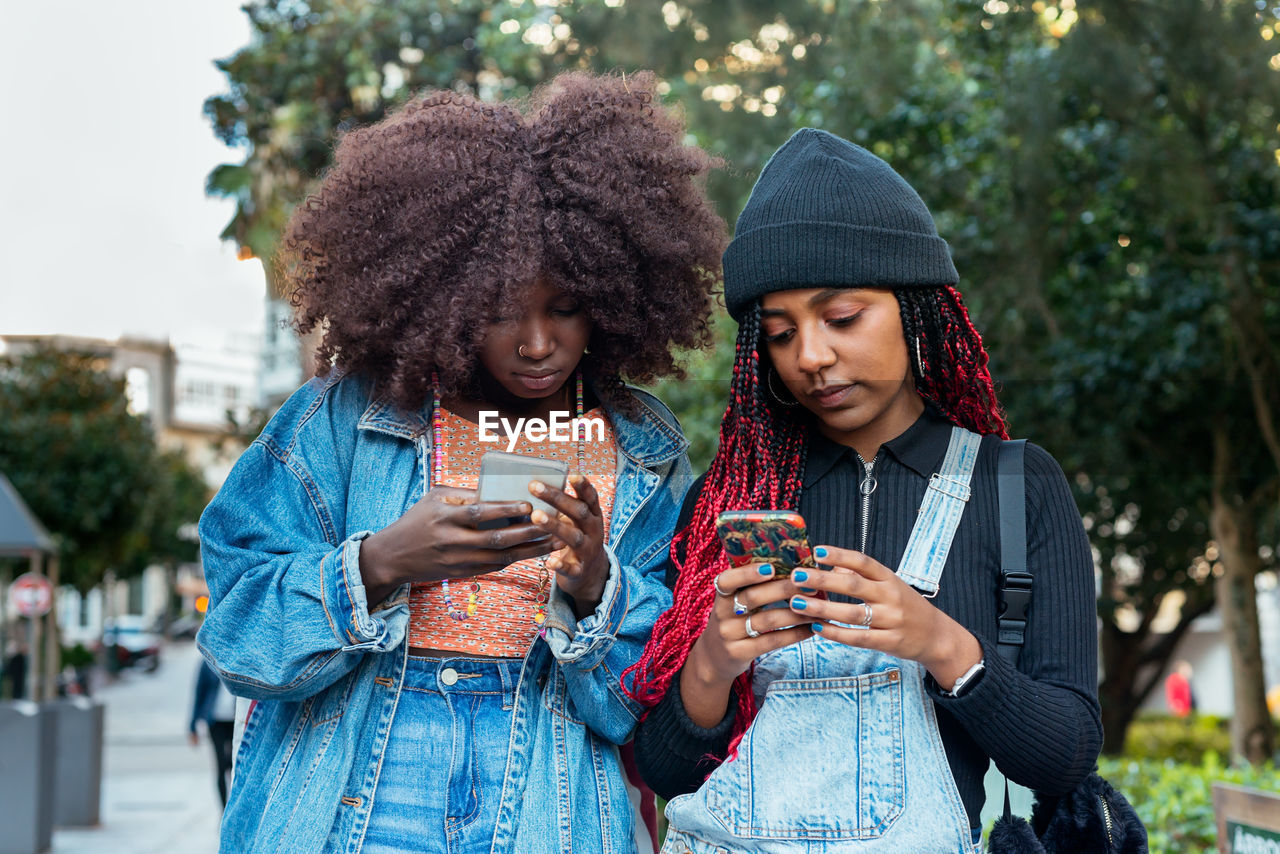 Young ethnic female friends with afro hairstyles concentrating while standing on blurred city street together and browsing mobile phones