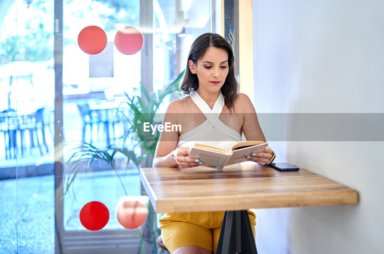 Attentive young female reading interesting story in book while sitting at table and spending weekend in cafe