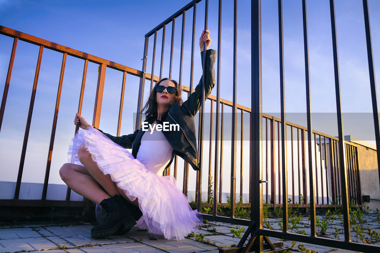 A ballerina in tutu sitting at the fence on the roof at sunset in boots and a jacket