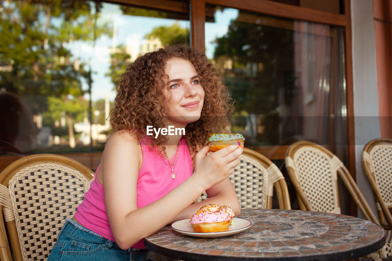 Portrait of beautiful curly young woman in cafe on summer terrace, delicious colorful donuts, 