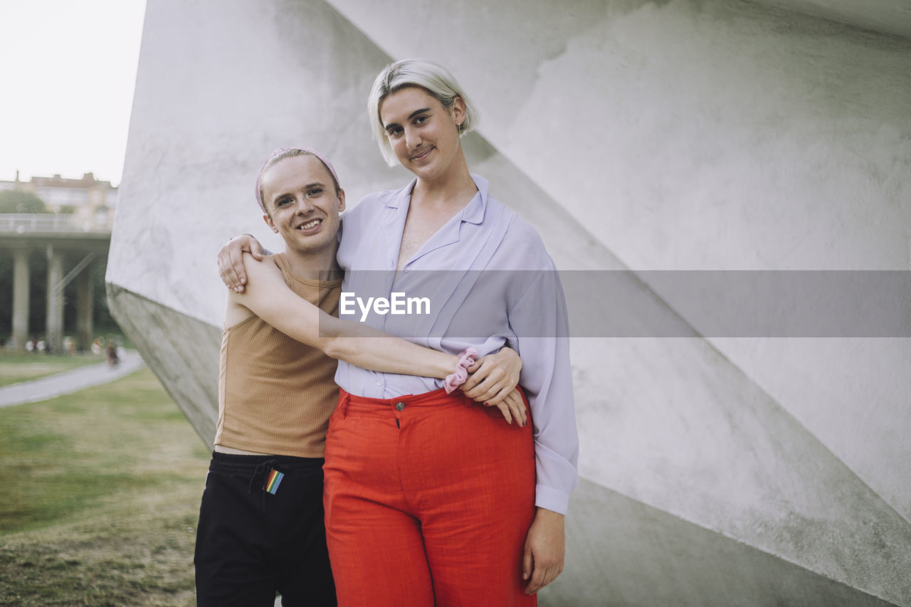 Portrait of smiling non-binary friends standing against wall at park