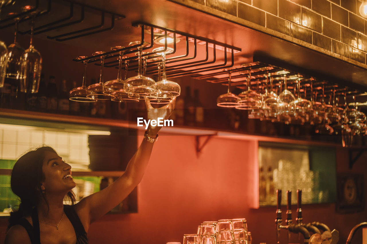 Smiling female bartender hanging cocktail glasses on rack while working at bar