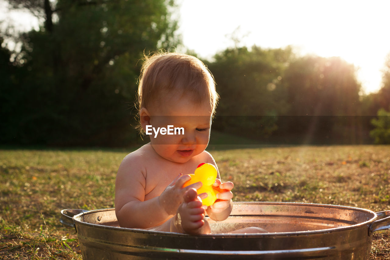 Cute little kid sits in a basin of water in nature and has rays of the setting sun, golden bathe. 