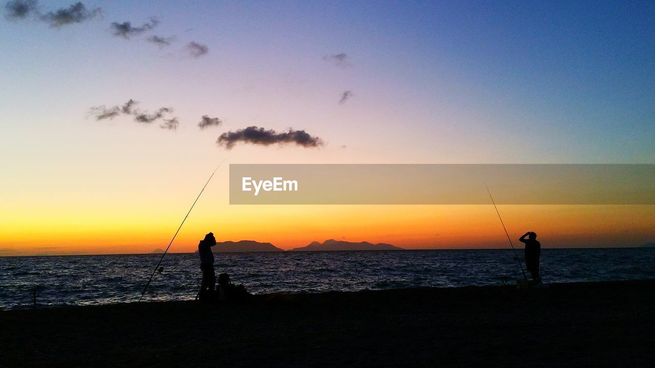 Silhouette men fishing on beach against sky during sunset