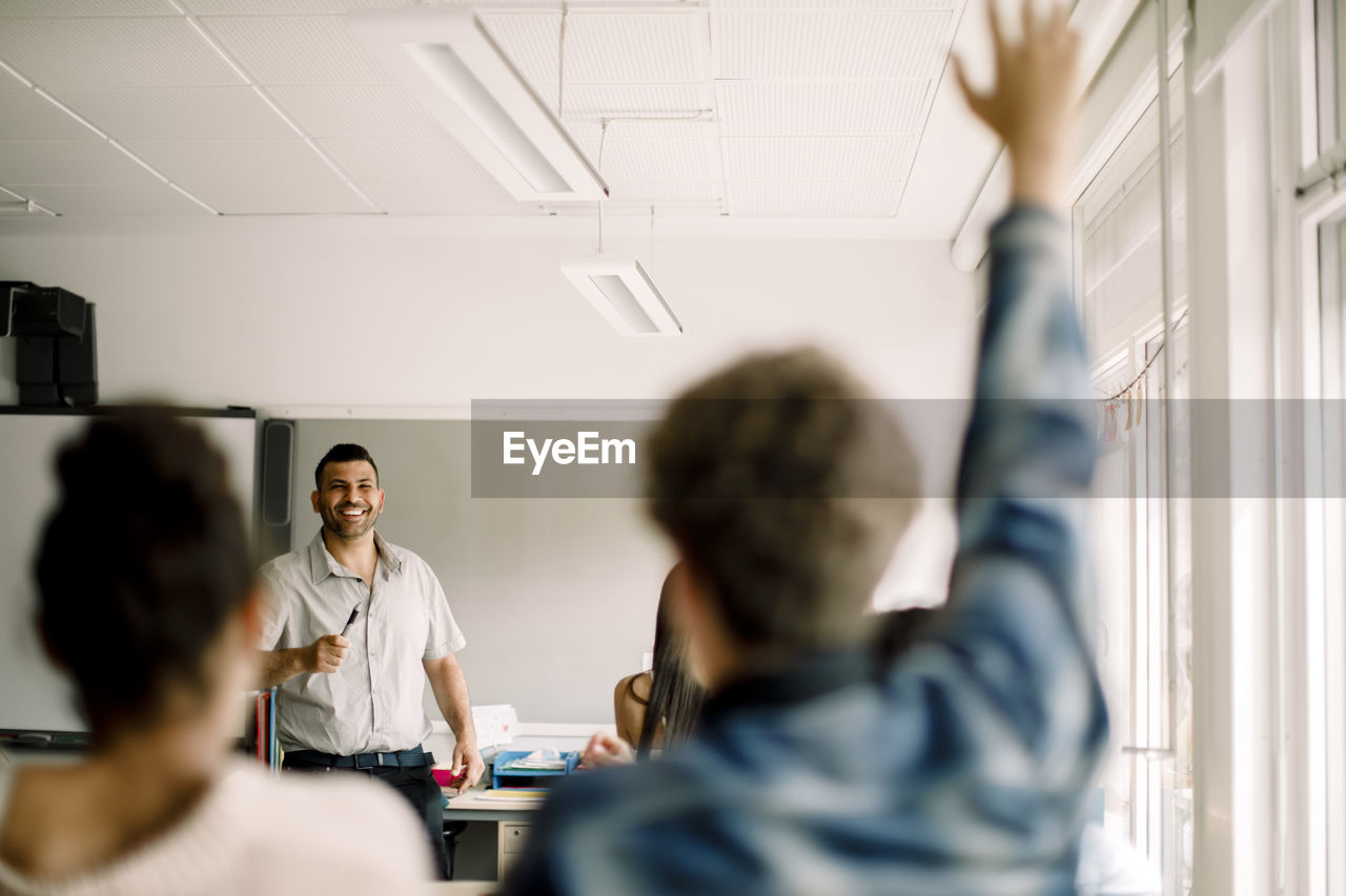 Male student with hand raised while smiling tutor standing in classroom