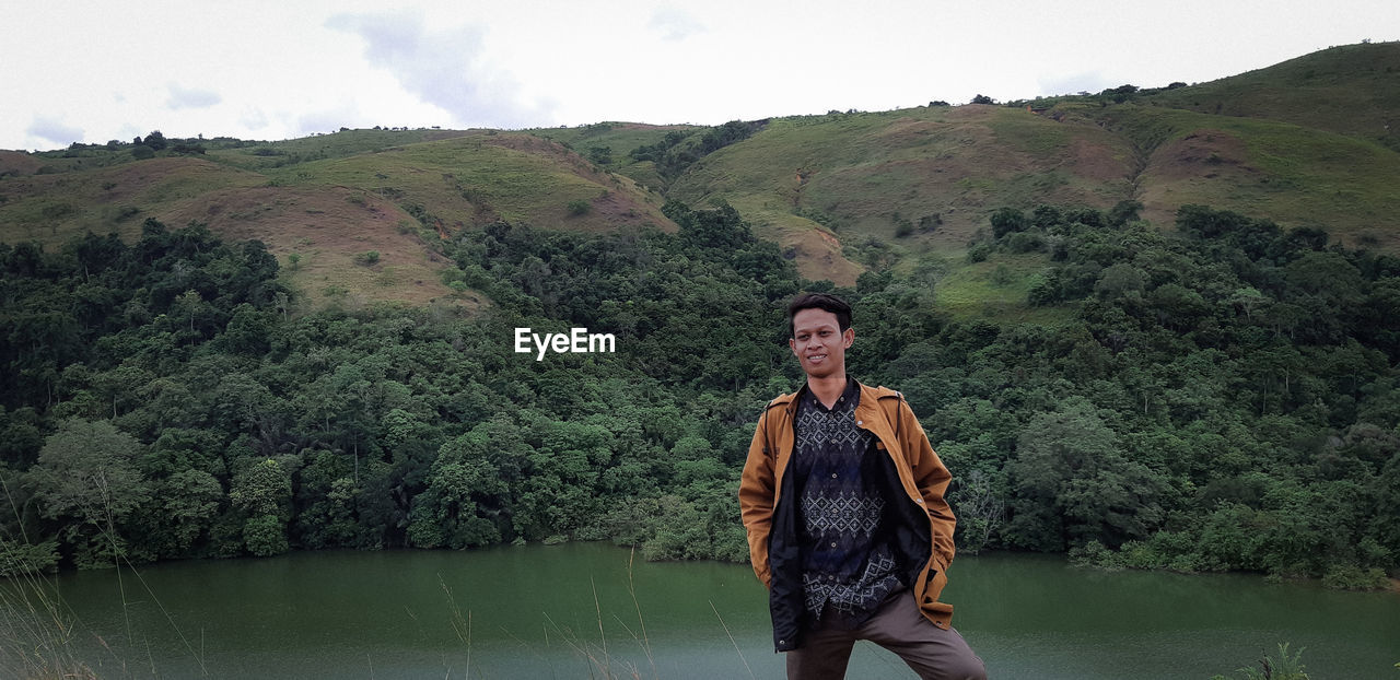 Smiling young man standing against river and mountain in forest