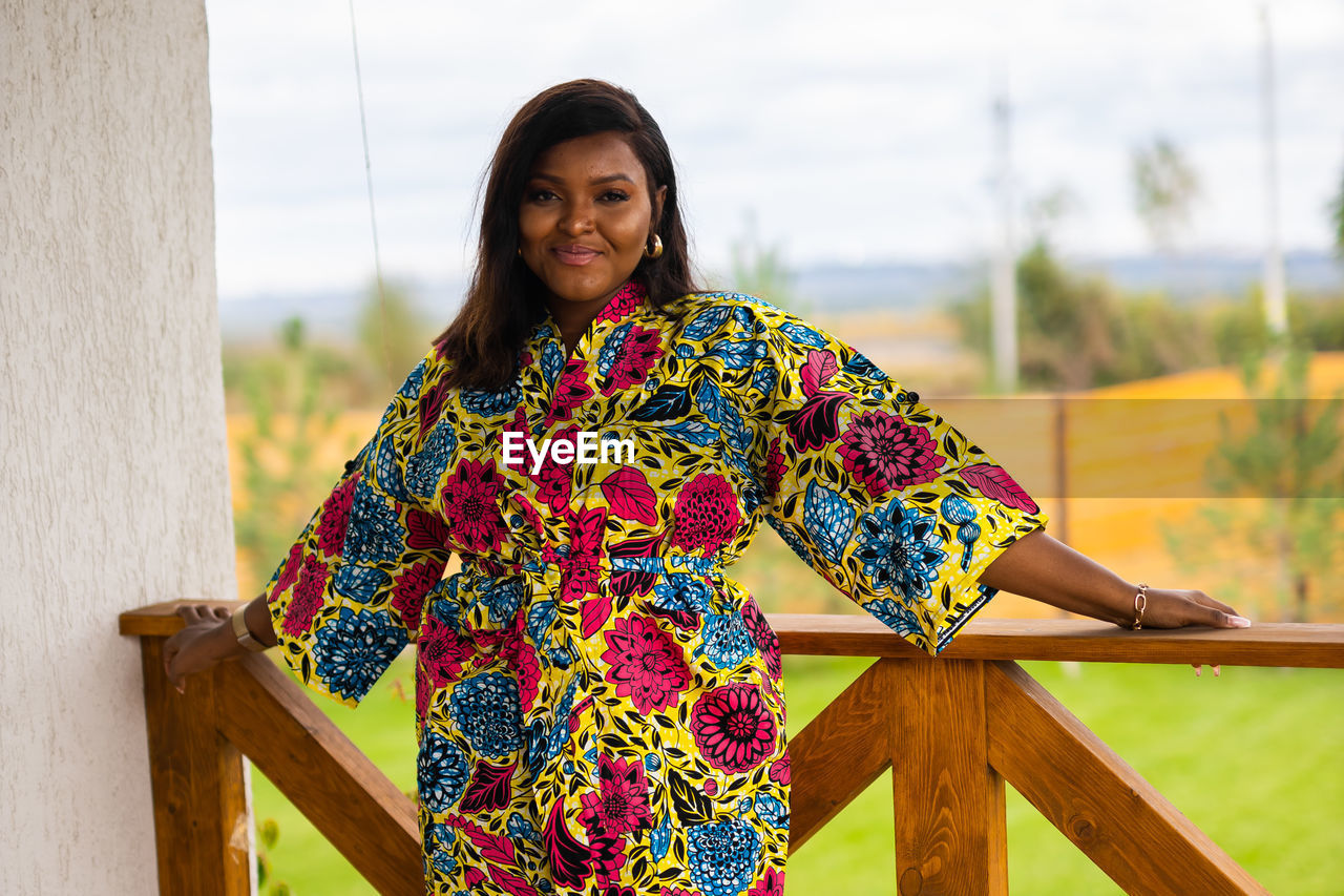 one person, spring, smiling, adult, women, yellow, portrait, happiness, looking at camera, dress, standing, clothing, three quarter length, front view, lifestyles, emotion, nature, hairstyle, young adult, cheerful, female, day, photo shoot, floral pattern, pattern, waist up, architecture, leisure activity, outdoors, focus on foreground, multi colored, wood, relaxation, traditional clothing, person, casual clothing, fashion, teeth, smile, brown hair, sky, enjoyment, long hair