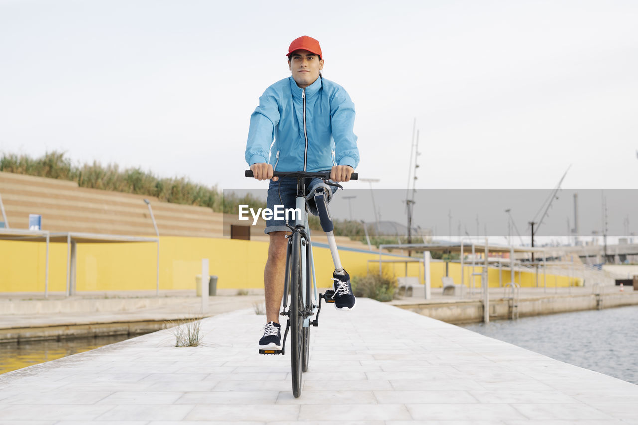 Young man with artificial limb and foot riding bicycle on footpath against clear sky