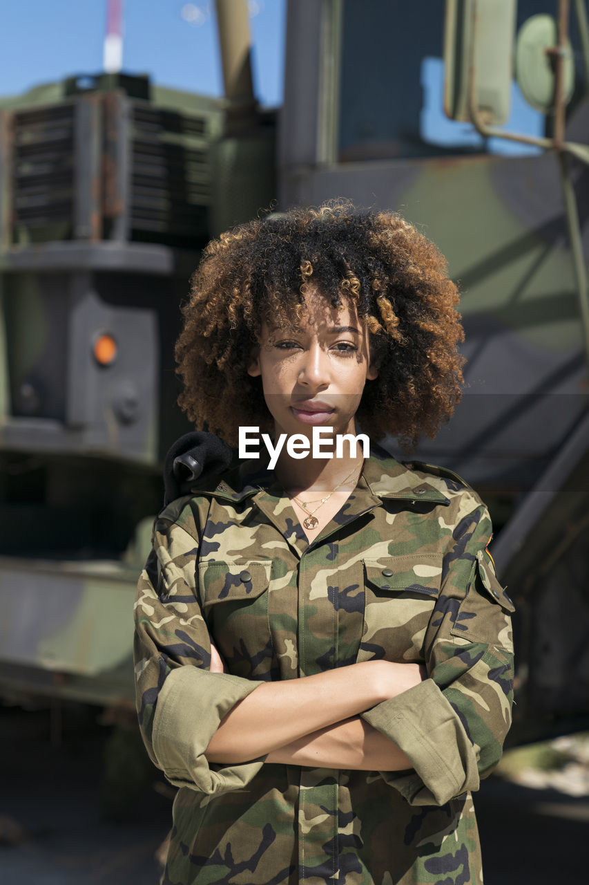 Confident young female army soldier with arms crossed standing against truck at military base