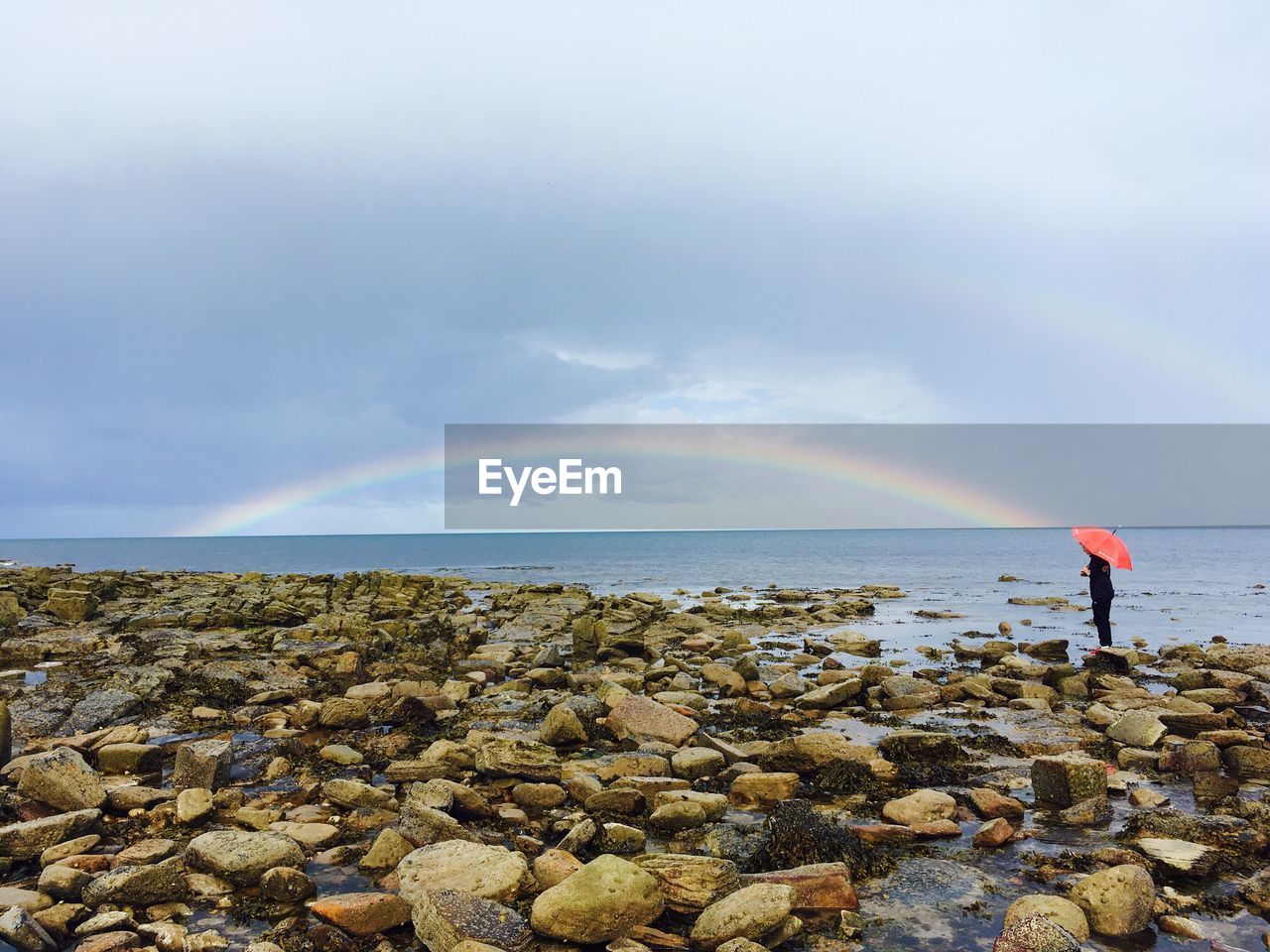 Person standing by sea with rainbow against sky during rain