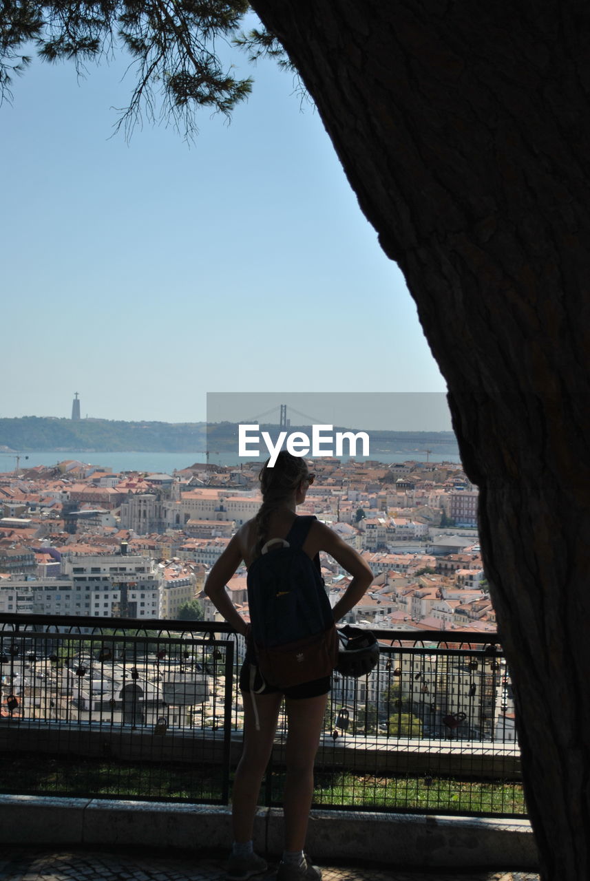 Rear view of woman with hand on hip standing against 25 de abril bridge