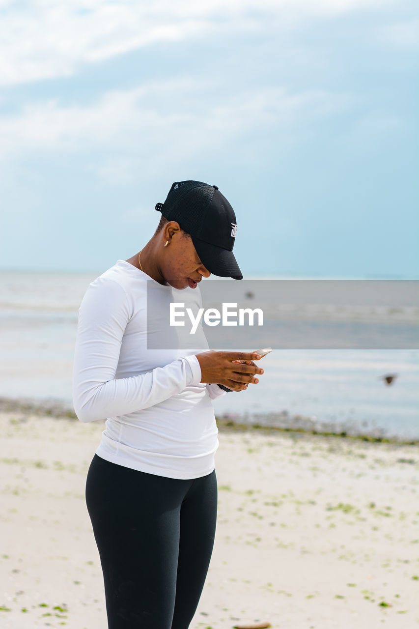 Side view of woman standing at beach looking at her smartphone against sky