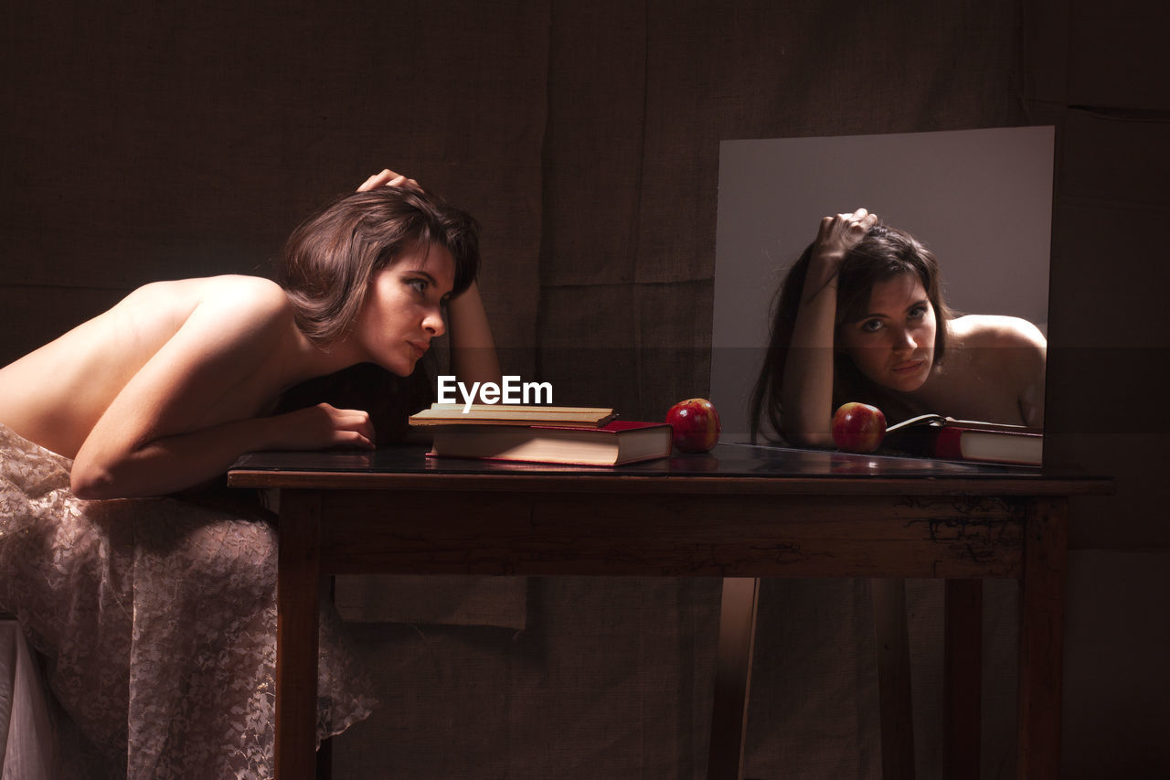 Shirtless young woman sitting on table while looking in mirror