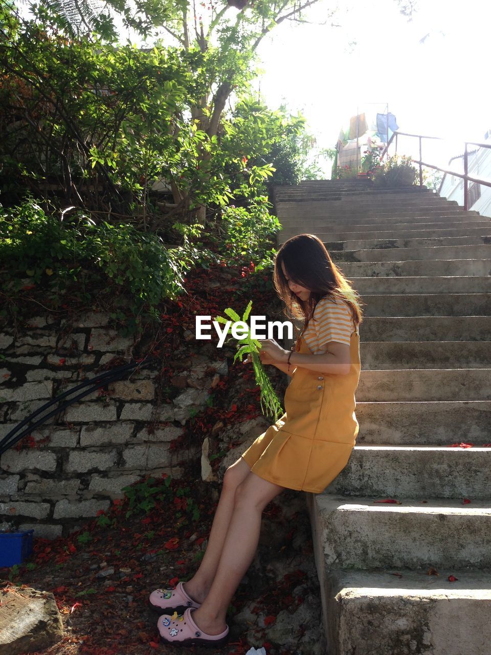 SIDE VIEW OF YOUNG WOMAN SITTING ON STAIRCASE AGAINST PLANTS