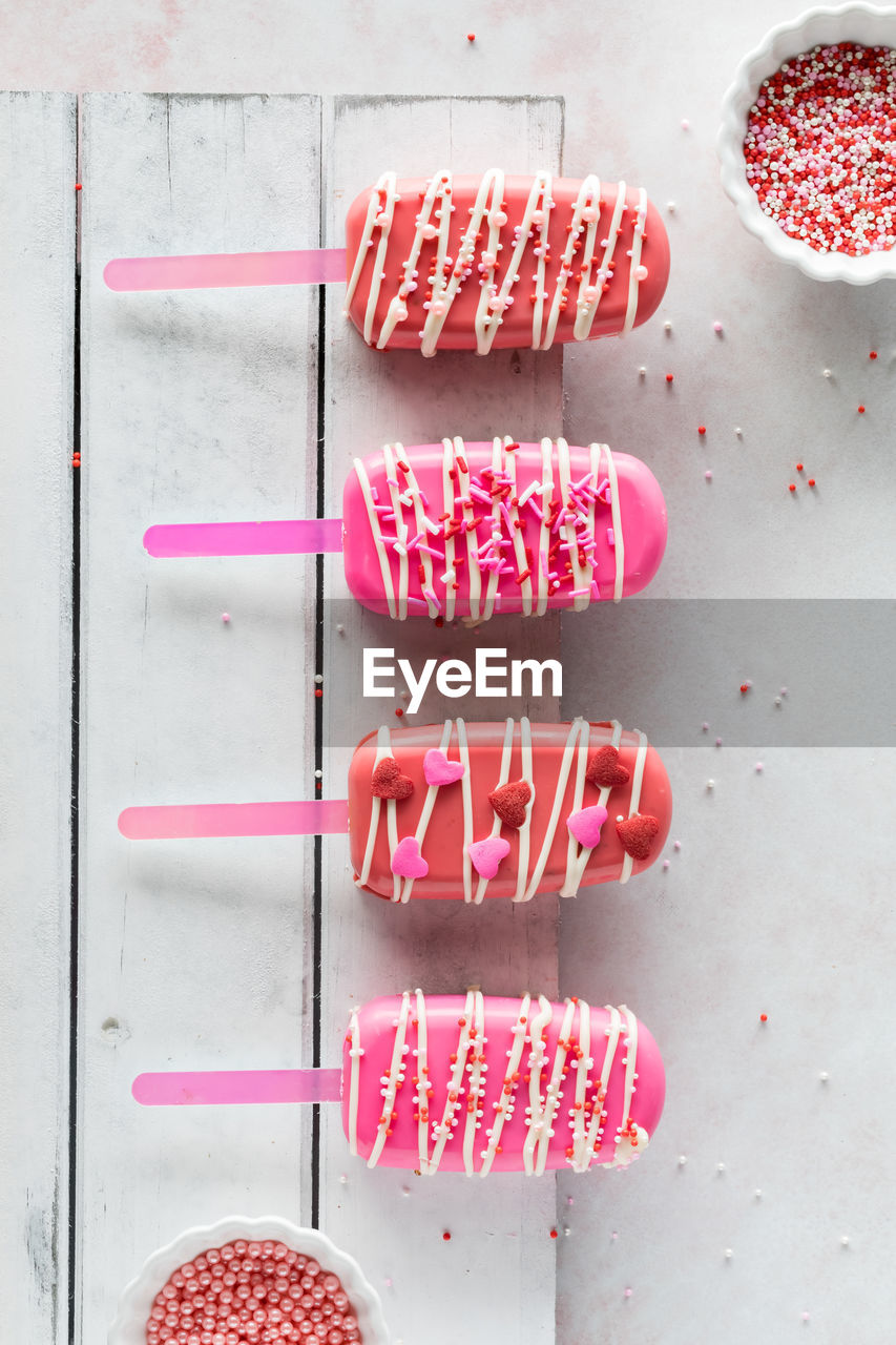 Homemade valentine cakesicles on a white rustic board with bowls of sprinkles.