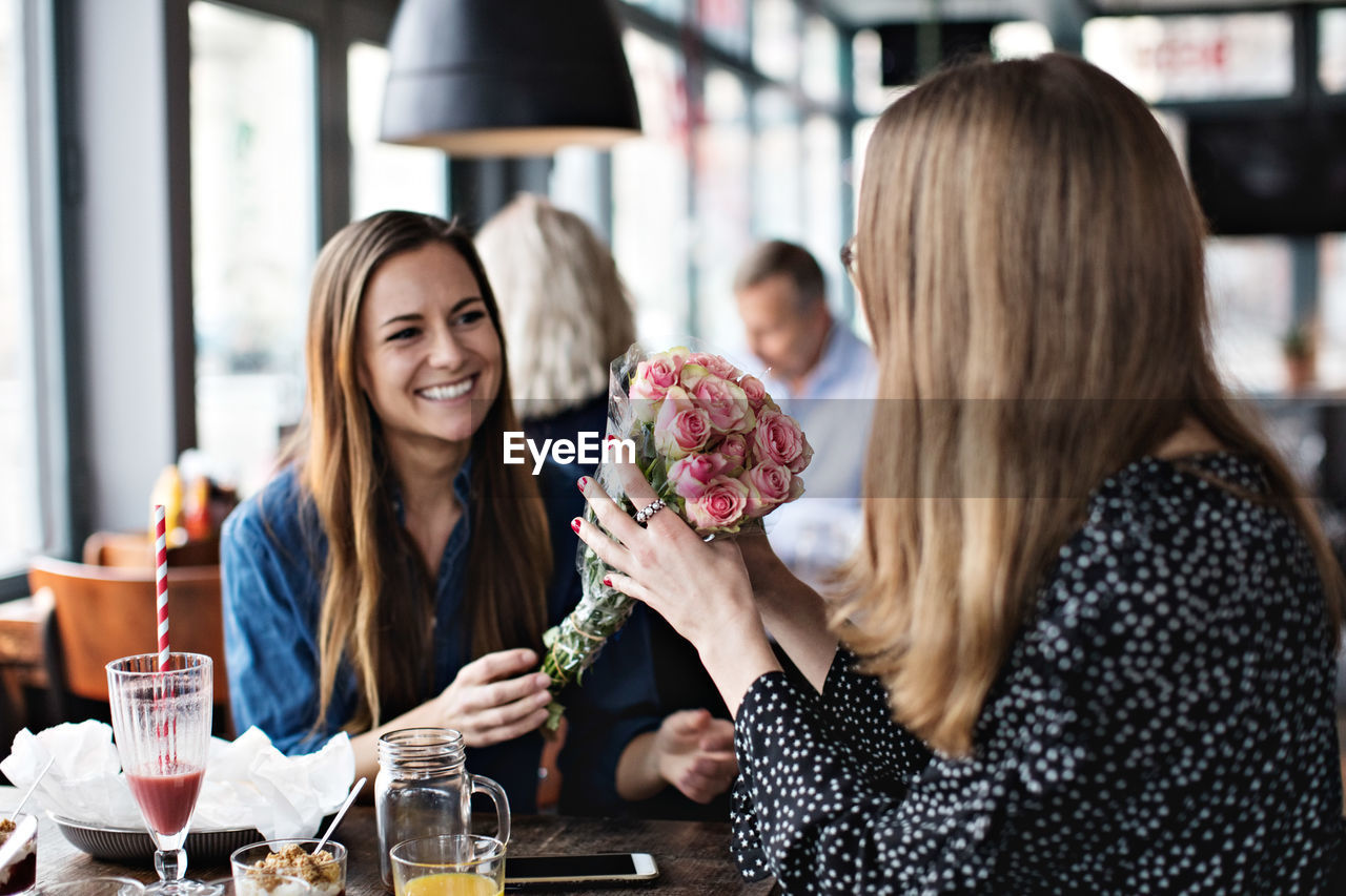 Smiling young woman giving fresh flower bouquet to female friend sitting at restaurant