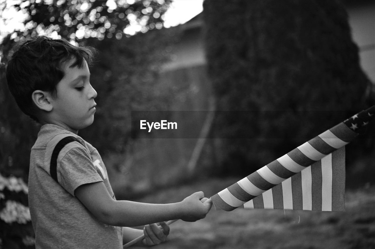 Side view of boy holding american flag while standing in yard