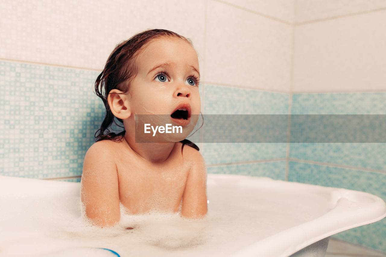 Topless girl with mouth open looking away while sitting in bathtub at home