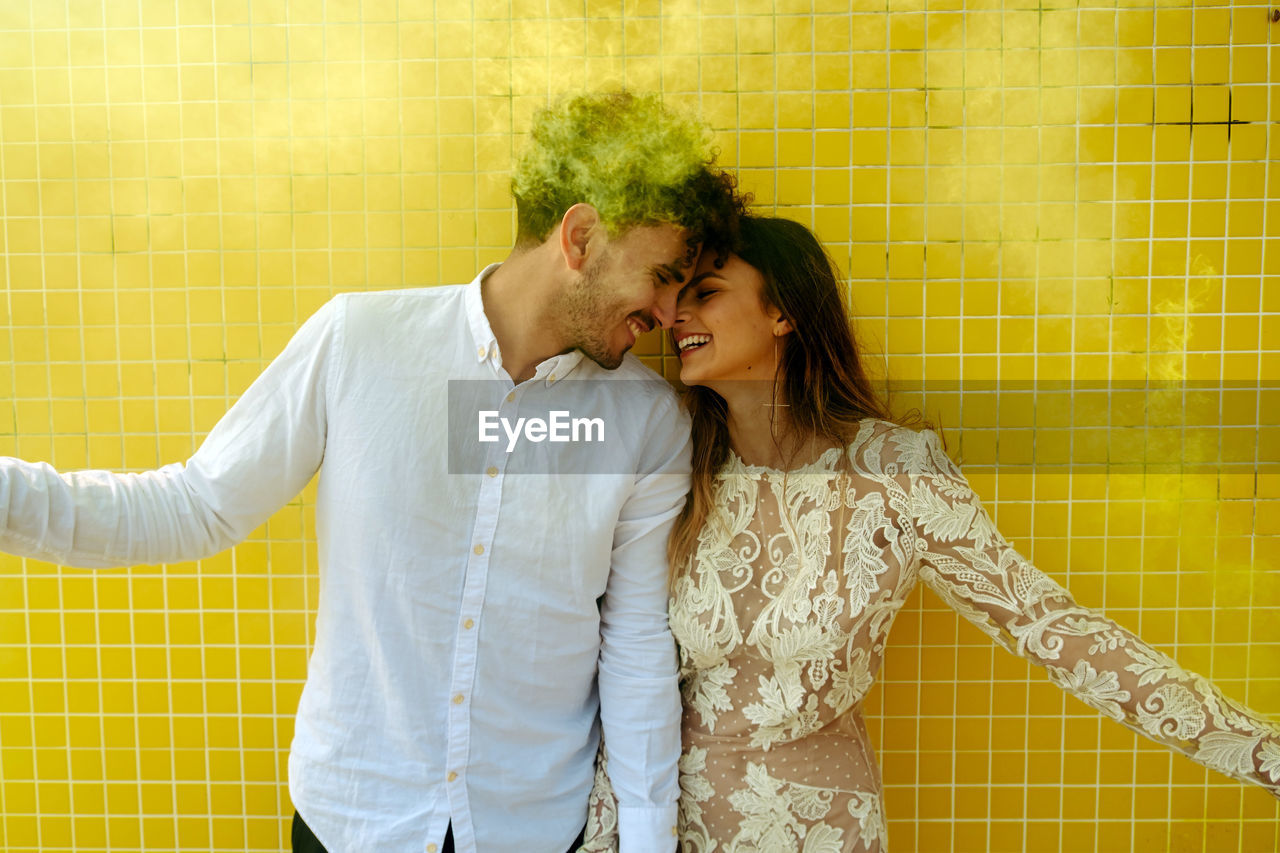 Young romantic newlywed couple in stylish wedding outfits having fun with yellow smoke bombs while standing together near modern building in tropical city