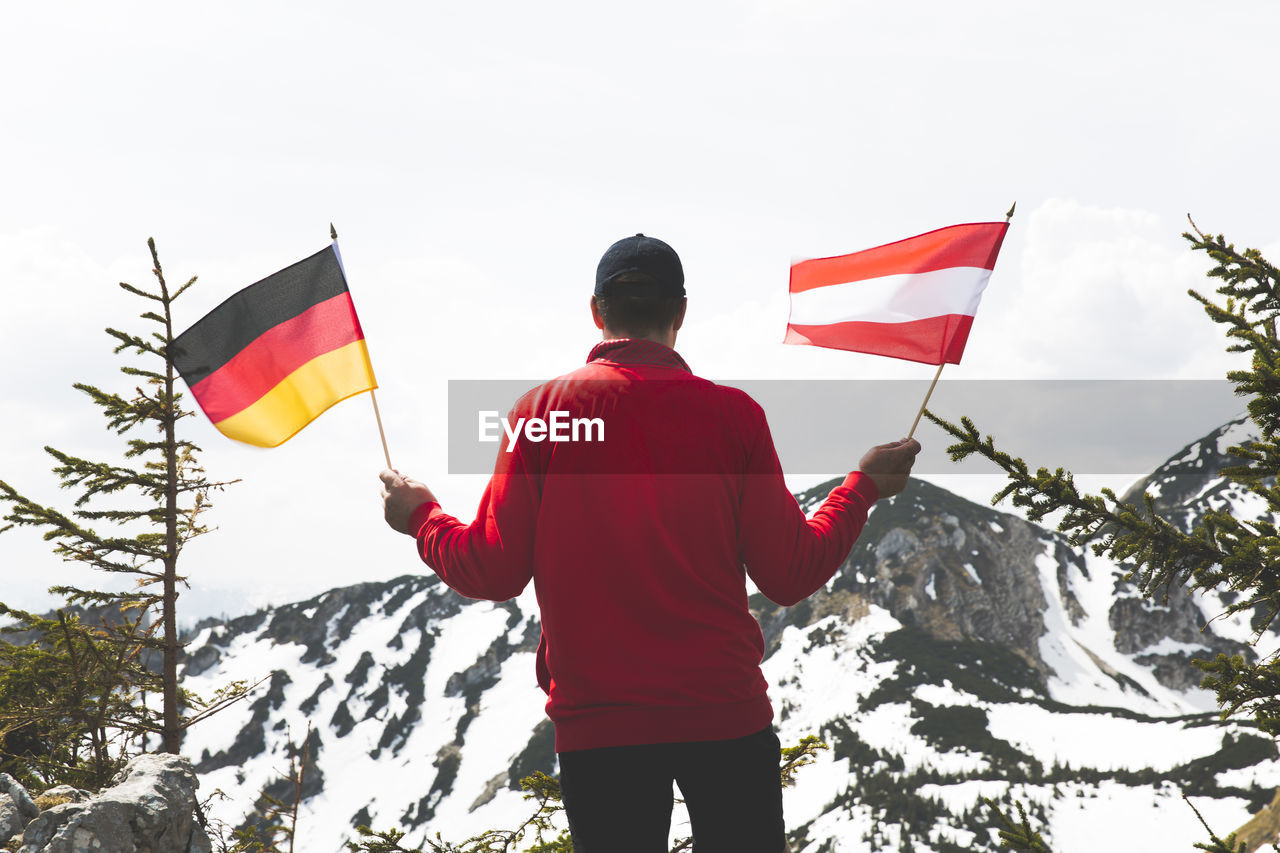 Rear view of mature man holding national flags while standing against snowcapped mountain