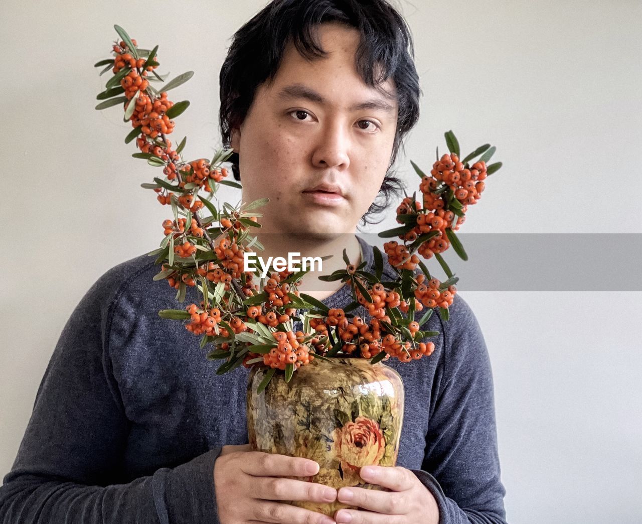 Young asian man holding arrangement of orange rowan berry branches in a vase.