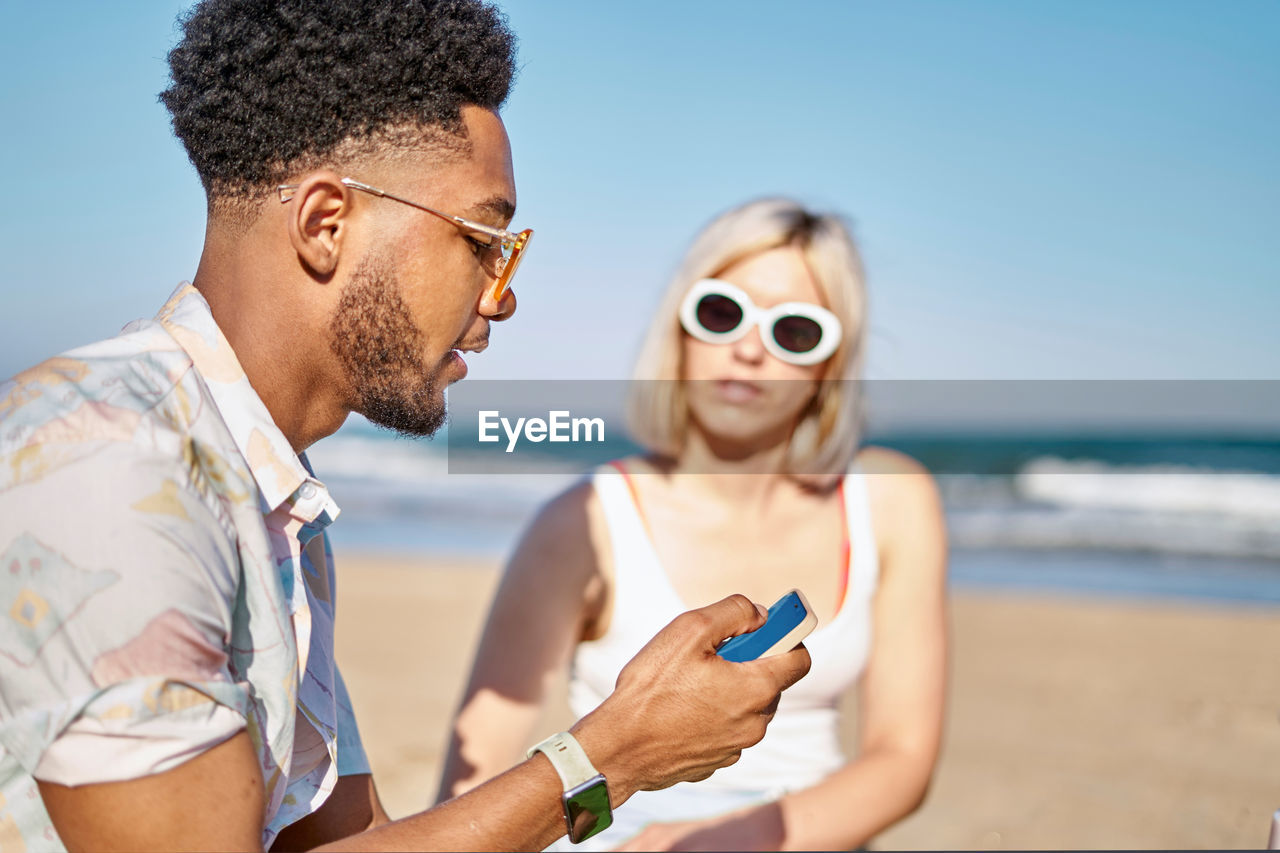 Black man sharing news in social media with girlfriend while spending time on sandy beach against sea and blue sky together