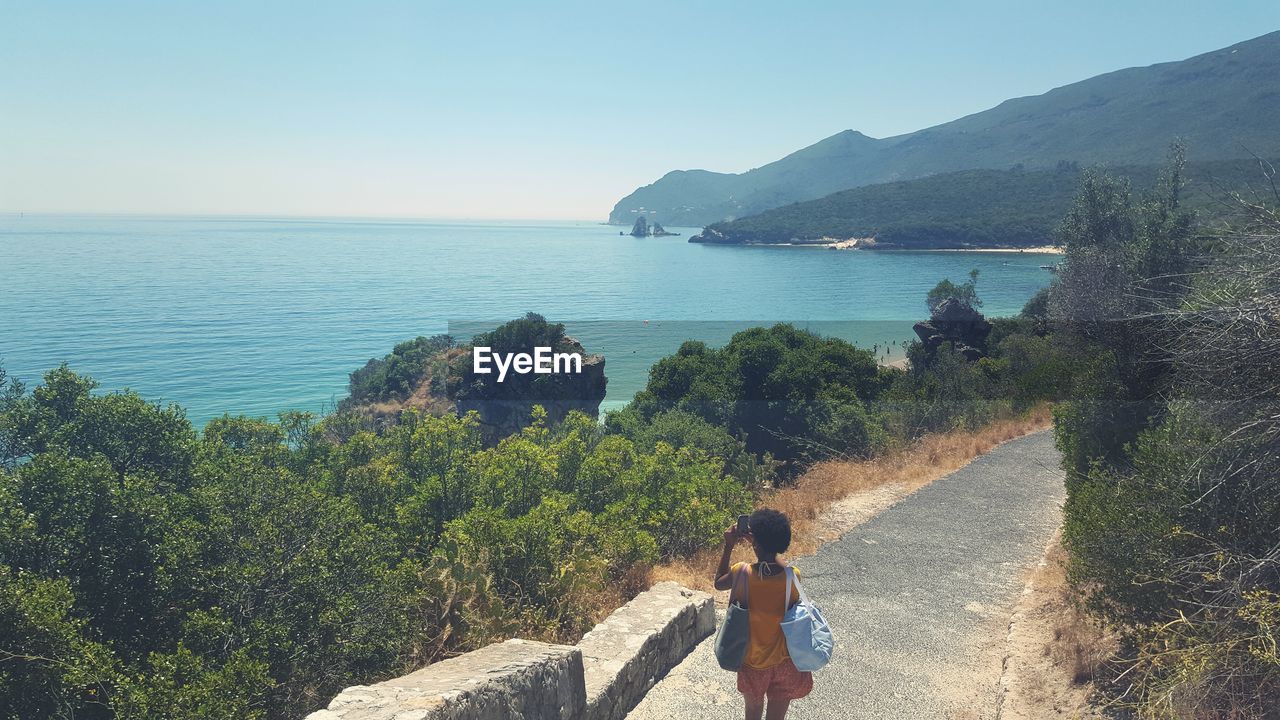 High angle view of woman standing on pathway amidst trees by sea against sky