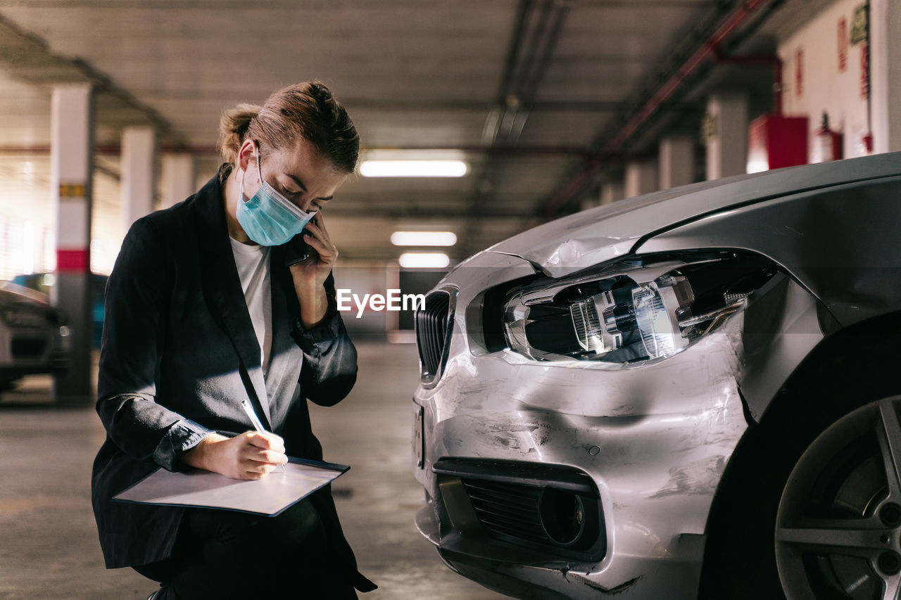 Unrecognizable concentrated female insurance officer in formal suit and protective mask talking on mobile phone while inspecting car damages and filling out claim after accident