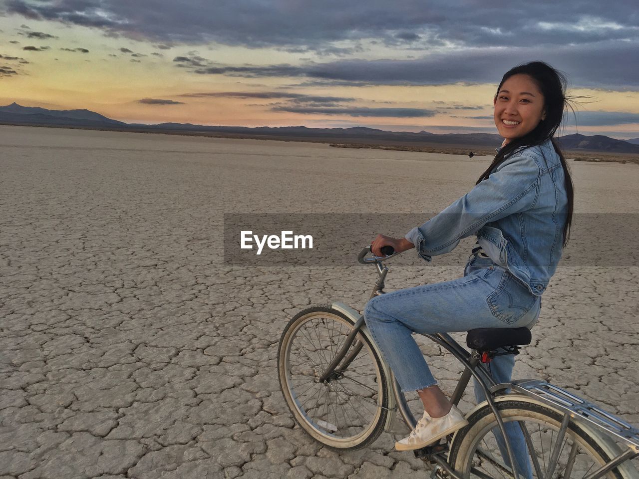 Portrait of smiling young woman riding bicycle on cracked field at sunset