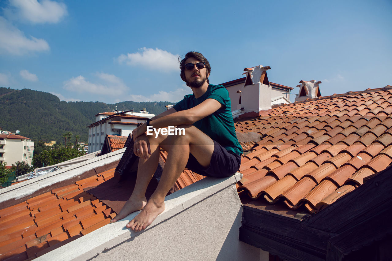 Man in the green t-shirt shorts and sunglasses sitting on the roof of a clay orange tile