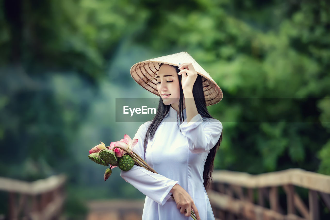 Woman wearing conical hat while holding flowers and pods against trees