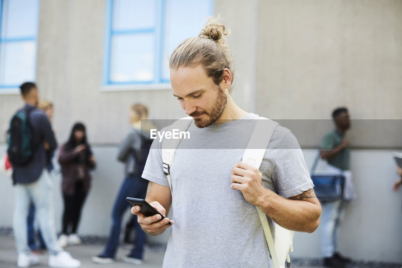 Male university student using mobile phone at campus with friends standing in background