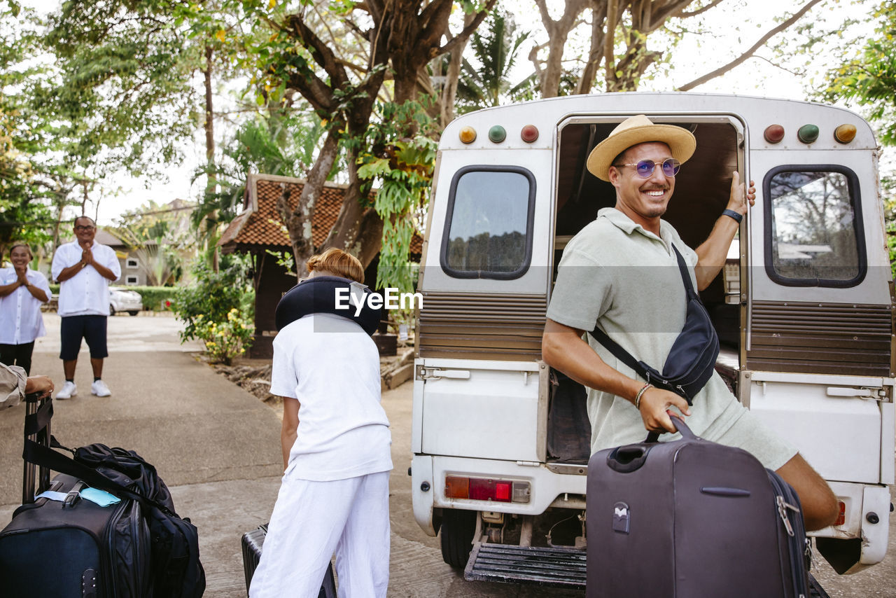 Smiling male tourist getting down with luggage from van at resort during vacation