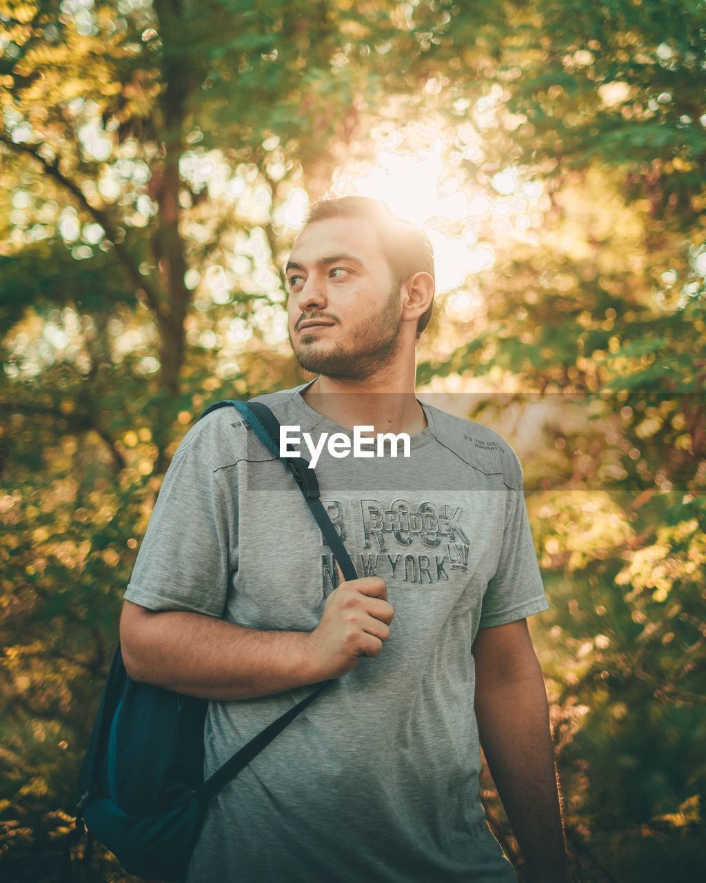 Young man looking away while standing against trees