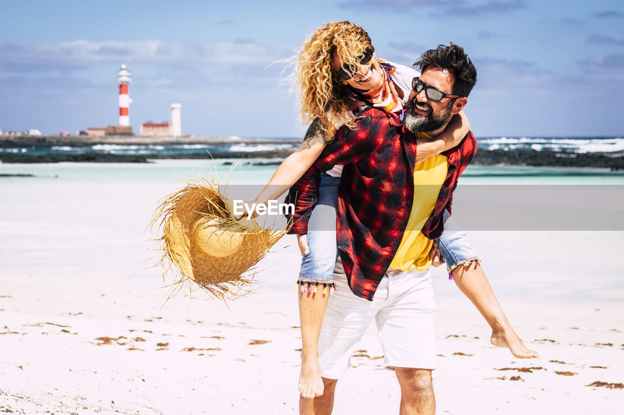 Cheerful man with woman piggy back standing at beach against sky