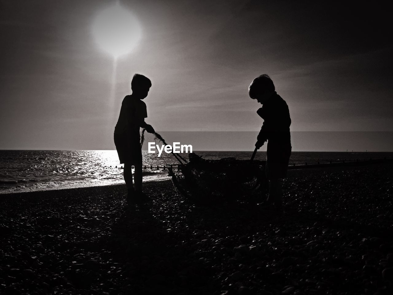 Silhouette boys pulling basket on beach against sky